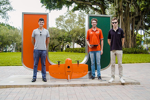 This is a photo of three University of Miami alumni. They are standing in front of the "U" statue. There is a drone on the ground in front of them. The photo was taken outside.